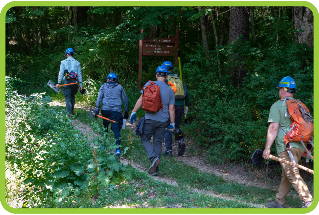 Volunteers walk with tools past forested sign during trail stewardship event