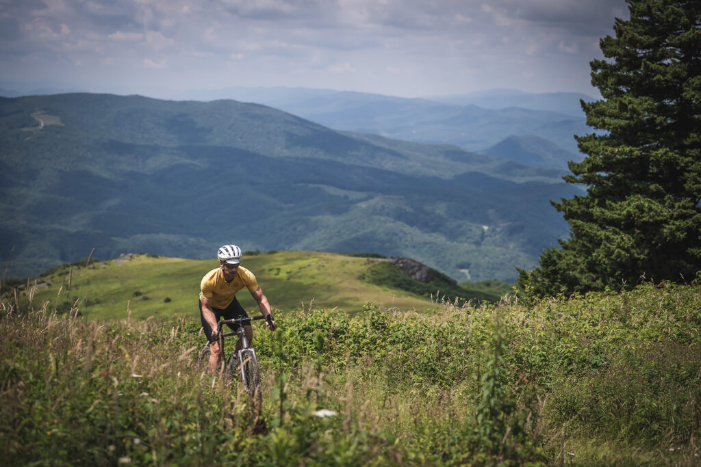 Biker summits gravel trail in grassy meadow with rolling green mountains in the background. 